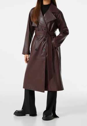 Vrouw die een lange donkerbruine leren trenchcoat met riem draagt, zwarte brede broekspijpen en zwarte stevige schoenen, staand tegen een witte achtergrond.