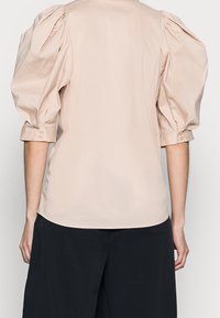 Beige blus med puffärmar och knäppta manschetter, bärs med svarta vida byxor, sedd bakifrån.