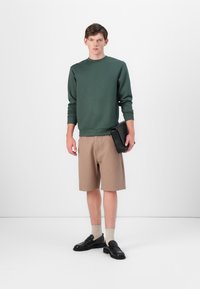 Sweatshirt verde de gola redonda, calções bege às riscas, sapatos pretos sem atacadores, meias crème, segurando uma clutch preta. Design simples e moderno.