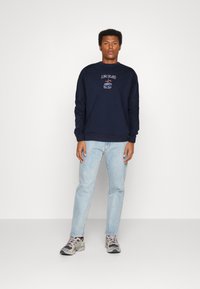 Sudadera de marina con el texto "LONG ISLAND", cuello redondo y diseño bordado. Combinada con jeans de color azul claro y zapatillas grises.