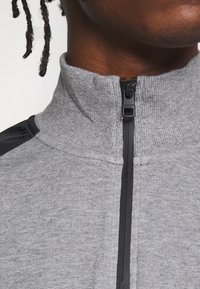Giacca grigia con zip e colletto alto, caratterizzata da un corpo in maglia testurizzata e pannelli neri sulle spalle. Include una cerniera con tirazip brandizzato.