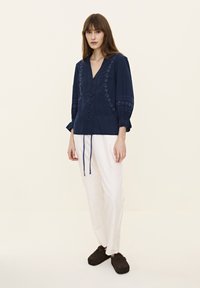 Navyblaue bestickte Bluse mit V-Ausschnitt, Knopfverschluss und Dreiviertelärmeln, kombiniert mit weißen, locker sitzenden Hosen und dunklen Clogs.
