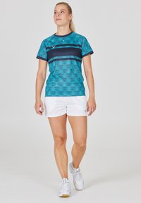 Träningsoutfit med en petrolfärgad mönstrad skjorta som har marinblå ränder och vita shorts. Skorna är ljusgrå med gula detaljer.