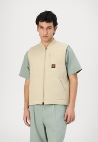 Beige ujjatlan mellény elöl cipzárral, két nagy zsebbel és egy logóval a mellkason, világoszöld, rövid ujjú ing felett viselve.