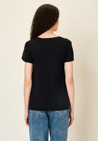 Woman with long curly hair wearing a plain black short-sleeve shirt and blue denim jeans, standing against a beige background.