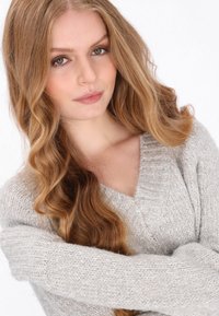 Pulover gros, de culoare gri deschis, cu decolteu în V și manșete nervurate. Textură moale și croială lejeră, purtat de un model cu părul lung și ondulat.