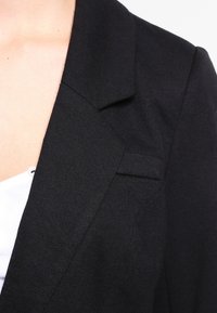 Černý blazer s klopovým límcem, strukturovanými rameny a jednou náprsní kapsou. Texturovaná látka s hladkým povrchem.