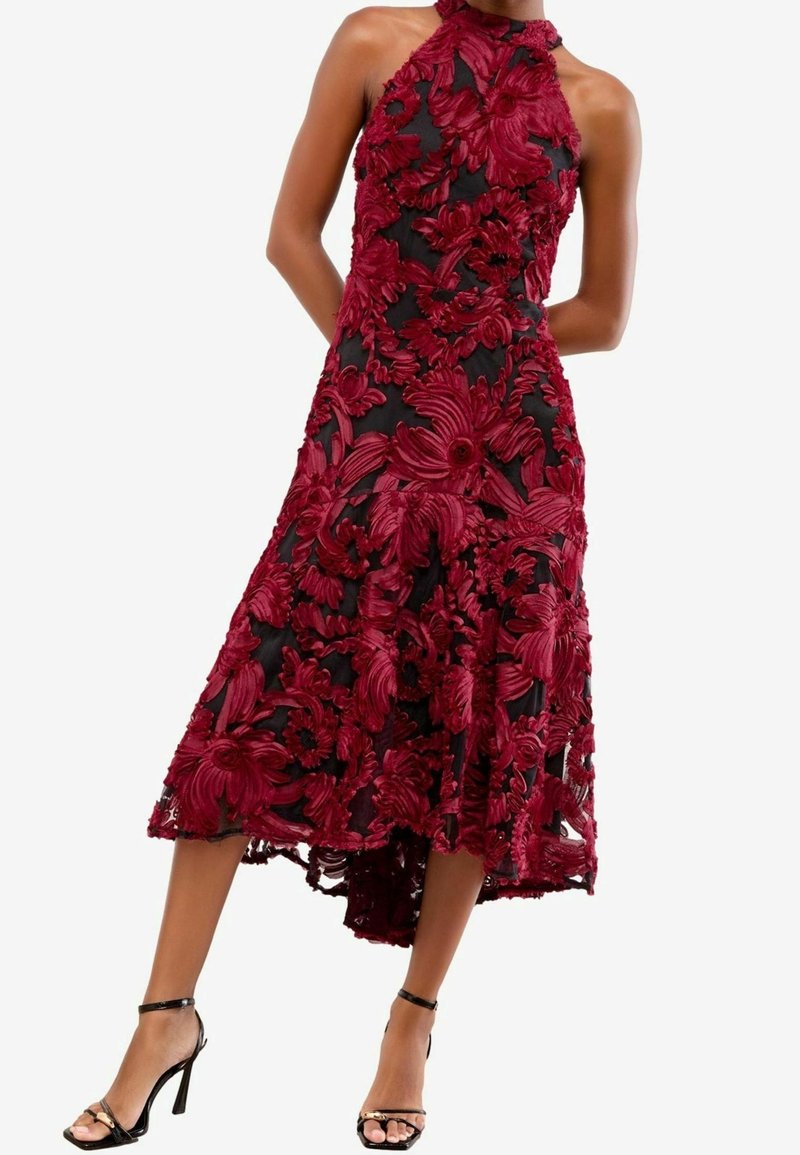 Robe noire sans manches avec broderie florale rouge foncé, encolure halter et jupe midi évasée, portée avec des sandales noires à talons hauts.