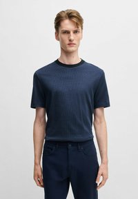 T-shirt blu navy realizzato in tessuto testurizzato, con colletto nero rotondo. Il modello indossa pantaloni blu scuro, in piedi davanti a uno sfondo neutro.