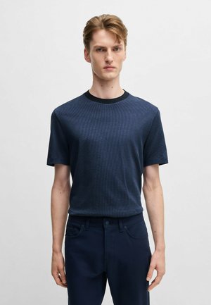 Marineblaue T-Shirt aus strukturiertem Stoff, mit einem runden schwarzen Halsausschnitt. Model trägt dunkelblaue Hose und steht vor einem neutralen Hintergrund.