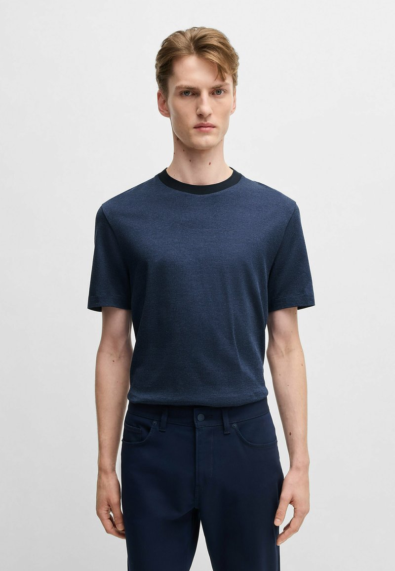 T-shirt blu navy realizzato in tessuto testurizzato, con colletto nero rotondo. Il modello indossa pantaloni blu scuro, in piedi davanti a uno sfondo neutro.