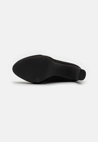 Zapato negro de tacón alto con parte superior de gamuza suave, punta redonda y suela de goma texturizada con estrías horizontales para mayor agarre.