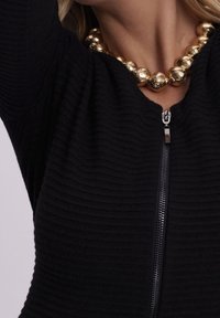 Indumento nero con zip e texture a coste, caratterizzato da una cerniera metallica e una collana di perle dorate grandi. Nessun dettaglio aggiuntivo sullo sfondo.