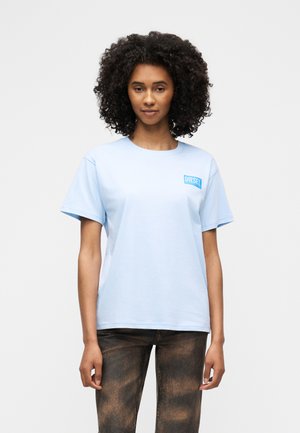 Frau mit lockigem Haar, die ein hellblaues Diesel-T-Shirt und dunkel gewaschene Jeans trägt, vor einem schlichten weißen Hintergrund stehend.