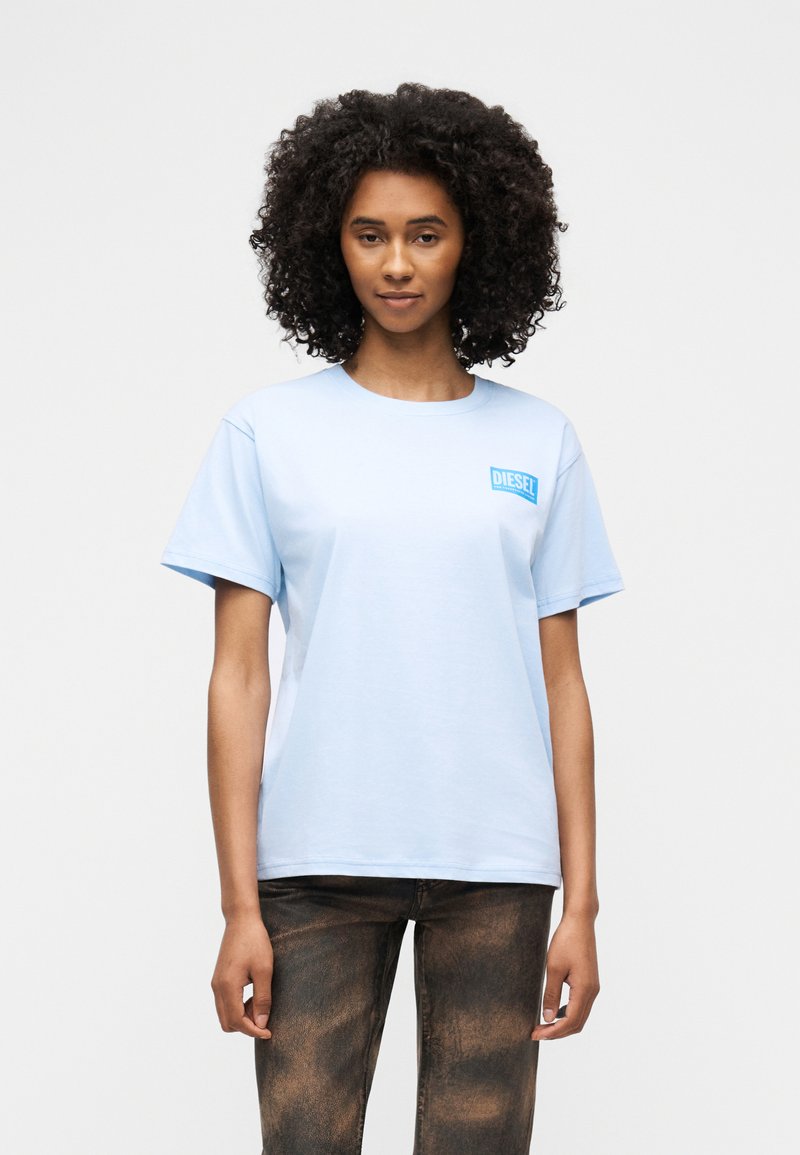 Γυναίκα με σγουρά μαλλιά, φορώντας ένα ανοιχτό μπλε t-shirt Diesel και σκούρα τζιν, στέκεται μπροστά σε απλό λευκό φόντο.