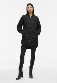 Γυναίκα φορώντας ένα μακρύ μαύρο επενδυμένο παλτό με κουμπιά, μαύρες skinny τζιν και μαύρες ψηλοτάκουνες μπότες μέχρι τον αστράγαλο, στέκεται με τα χέρια στην τσέπη.