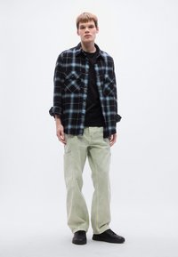 Καρό φανελένιο πουκάμισο σε μαύρο και τυρκουάζ, φορεμένο πάνω από μαύρο μπλουζάκι. Φαρδιά παντελόνια cargo σε ανοιχτό πράσινο με πλαϊνές τσέπες και μαύρα αθλητικά παπούτσια.