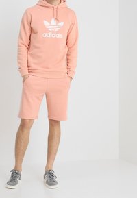 Ljusrosa sweatshirt med vit Adidas-logga, ihop med matchande shorts. Modellen bär grå sneakers. Slät textur, avslappnad design.