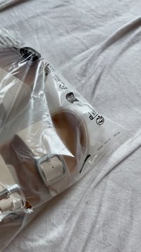 Il packaging di plastica contiene un oggetto beige chiaro con una fibbia rettangolare argentata e una cinturino morbido. Lo sfondo è in tessuto bianco.