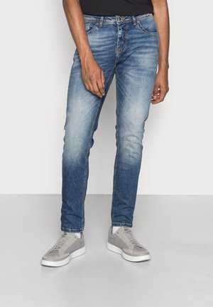 Blauwe denim jeans met een slim fit, een vervaagde wassing en subtiele stikseldetails, gecombineerd met grijze sneakers op een witte achtergrond.