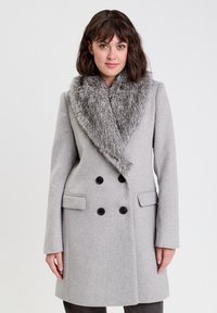 Abrigo gris de doble botonadura con un gran cuello de piel sintética, bolsillos laterales y botones negros. Textura suave similar a la lana.