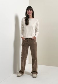 Femme debout contre un mur bicolore, portant une chemise blanche à manches longues, un pantalon large marron et des baskets blanches, les mains dans les poches, souriante.