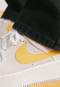 Sneakers Nike z białą zamszową cholewką, żółtym skórzanym logo swoosh, białymi sznurówkami i teksturowaną gumową podeszwą. W połączeniu z czarnymi dżinsami.
