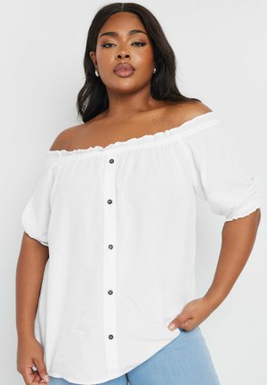 Witte off-shoulder bloes met ruches bij de halslijn, korte pofmouwen en zwarte knoopdetails aan de voorkant op een lichte achtergrond.
