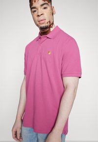 Polo de algodón rosa con cuello tipo polo, mangas cortas y un pequeño logo dorado en el pecho. Presenta un dobladillo recto y aberturas laterales.