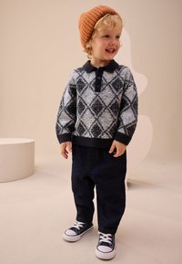 Jersey de punto con patrón de diamantes en gris y azul marino, cuello negro, tapeta de botones; pantalones de mezclilla oscuros; zapatillas azul marino; gorro de canalé naranja.