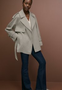 Plašč z vzorcem houndstooth v črni in beli barvi, z širokimi ovratniki, ožjim pasom in dolgimi rokavi, v kombinaciji s temnimi širokimi jeans hlačami.