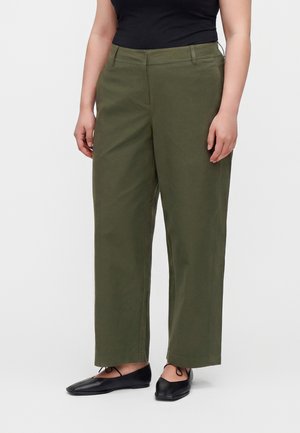 Femme portant un pantalon large couleur olive avec un haut noir sans manches et des chaussures plates noires, debout devant un fond uni.