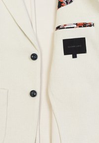 Blazer color crema con finitura testurizzata, dotato di bottoni neri, rivestimento del rever con motivo floreale e una etichetta di tasca nascosta.