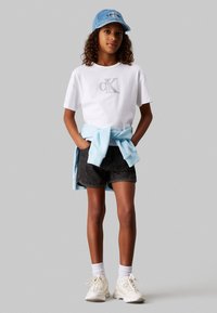 T-shirt bianca con ricamo "CK", pantaloni shorts in denim neri, felpa leggera blu chiaro legata in vita, cappellino di denim, sneakers bianche e calze bianche.