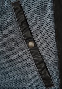 Texturerat grått tyg med ett meshnätmönster, med ett svart foder och en synlig rund knappdetalj på framsidan.