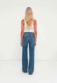 Mulher com cabelo loiro comprido, vestindo uma blusa branca sem mangas e calças jeans largas de cintura alta azuis, apoiada contra um fundo branco liso.