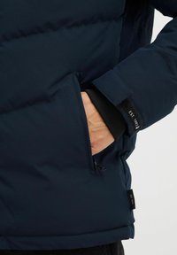 Chaqueta tipo puffer en color azul marino con un bolsillo con cremallera, puños acanalados y una etiqueta cosida en la manga. Material suave e impermeable con aislamiento.