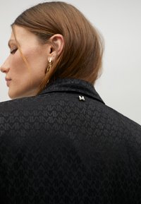 Černá bunda s texturovaným vzorem a límcem. Významný detail: malý zlatý akcent "M" poblíž límce.