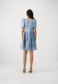 Robe bleue à volants avec des manches bouffantes, présentant un tissu texturé et des détails floraux. Portée avec des chaussures à talons hauts beiges.