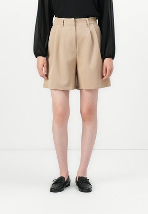 Kvinne iført beige skreddersydde shorts med folder, en svart langermet bluse og svarte loafers, står mot en hvit bakgrunn.