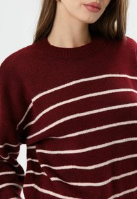 Een bordeauxrode trui met witte horizontale strepen, gemaakt van zacht materiaal, met een geribde ronde halslijn en een licht losse pasvorm.