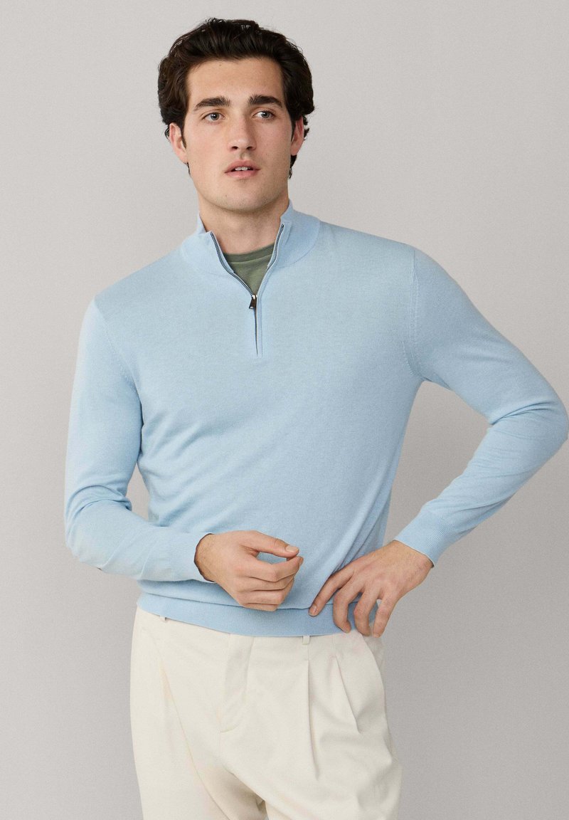 Homme portant un pull à quart de fermeture éclair bleu clair sur une chemise verte et un pantalon plissé beige, debout devant un fond uni.