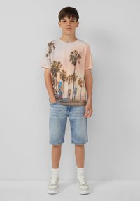 Koszulka graficzna z palmami w pastelowych kolorach, w zestawieniu z jasnymi dżinsowymi szortami. Białe sneakersy i prążkowane skarpetki sięgające kostek dopełniają stylizację.