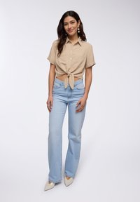Camicia beige a maniche corte legata in vita, abbinata a jeans a vita alta di colore blu chiaro e tacchi bianchi appuntiti, che mostrano una vestibilità rilassata.