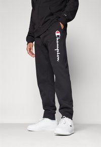 Svarte joggebukser med hvit "Champion" logo langs venstre ben, laget av myk stoff, kombinert med hvite sneakers, mot en ensfarget bakgrunn.