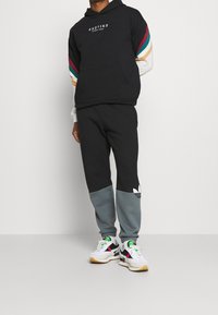 Felpa nera con maniche a blocchi di colore in teal, rosso e crema. Pantaloni della tuta neri con dettagli grigi e logo Adidas. Sneakers bianche e nere.