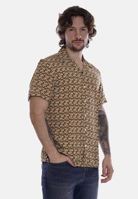 Camicia a maniche corte di colore giallo con un motivo circolare in tonalità di marrone e crema, dotata di chiusura a bottoni e vestibilità rilassata.