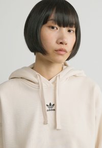 Sweatshirt bege com capuz feito de tecido macio, com bolso canguru. Apresenta o logotipo preto da Adidas acima do bolso. Textura suave, ajuste casual.