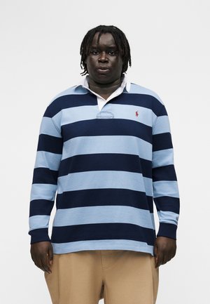 Uomo con pelle scura e dreadlocks che indossa una maglia da rugby a maniche lunghe a strisce blu navy e azzurro chiaro e pantaloni beige su sfondo bianco.