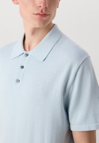 Svetlo modrá polo tričko vyrobené z mäkkej tkaniny. Obsahuje klasický golier, zapínanie na tri gombíky a malé vyšívané logo na hrudi.
